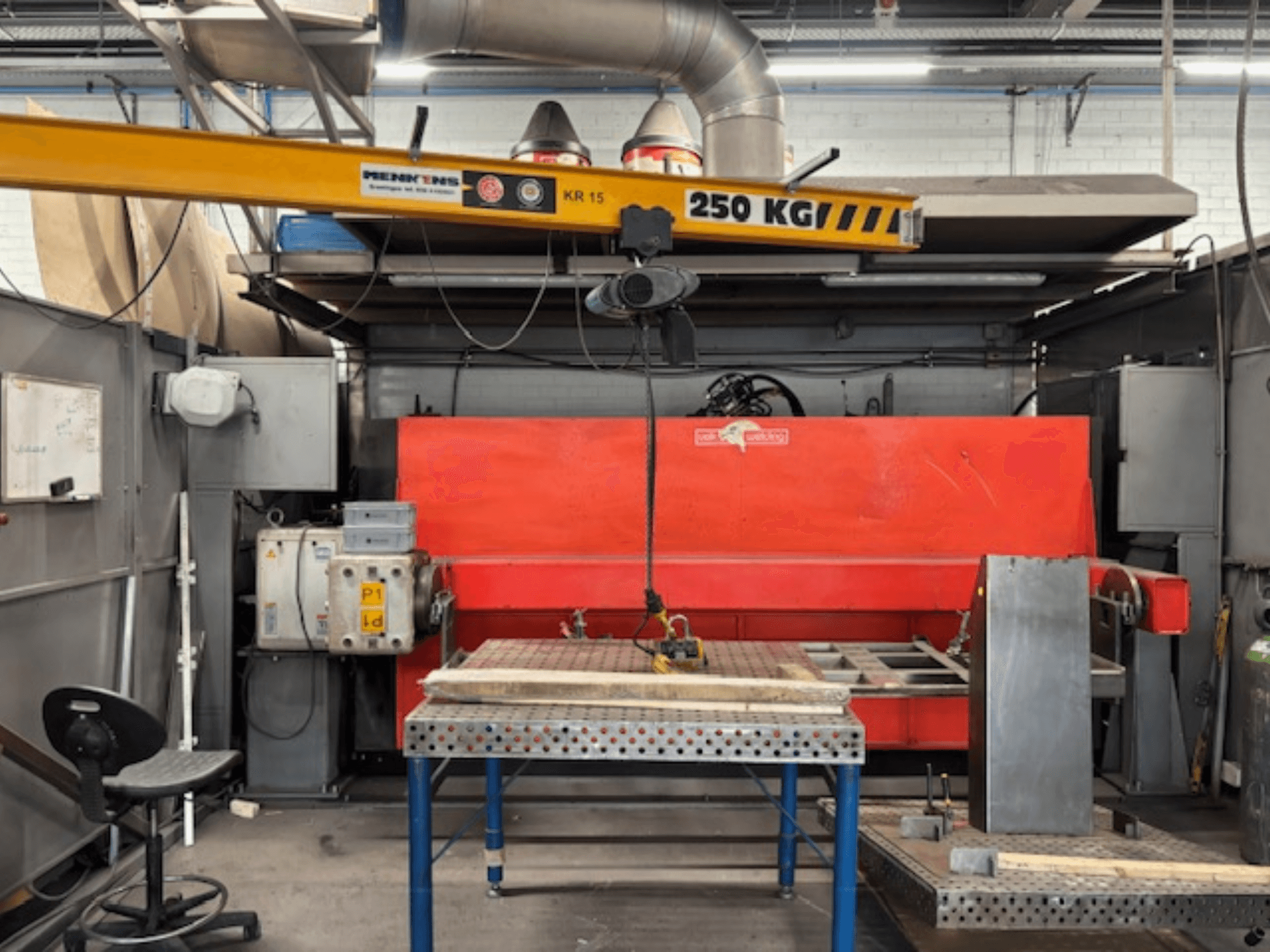 Industriverkstad med röd hydraulisk press, traverskran märkt "KR 15" och arbetsbord i metall, perspektiv från sidan.