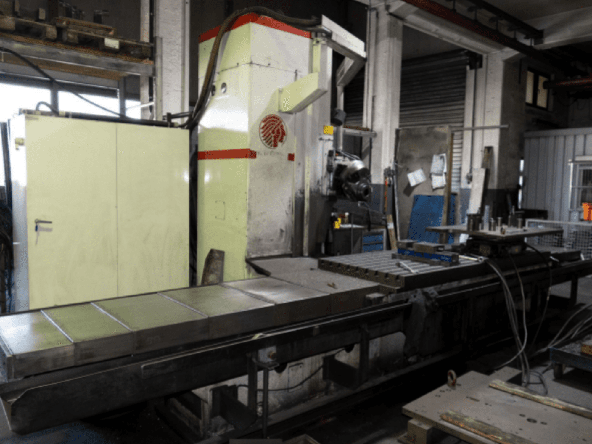 Industriell fräsmaskin med ett stort bord, sidovy, som visar reglage och tillbehör i en verkstadsmiljö.