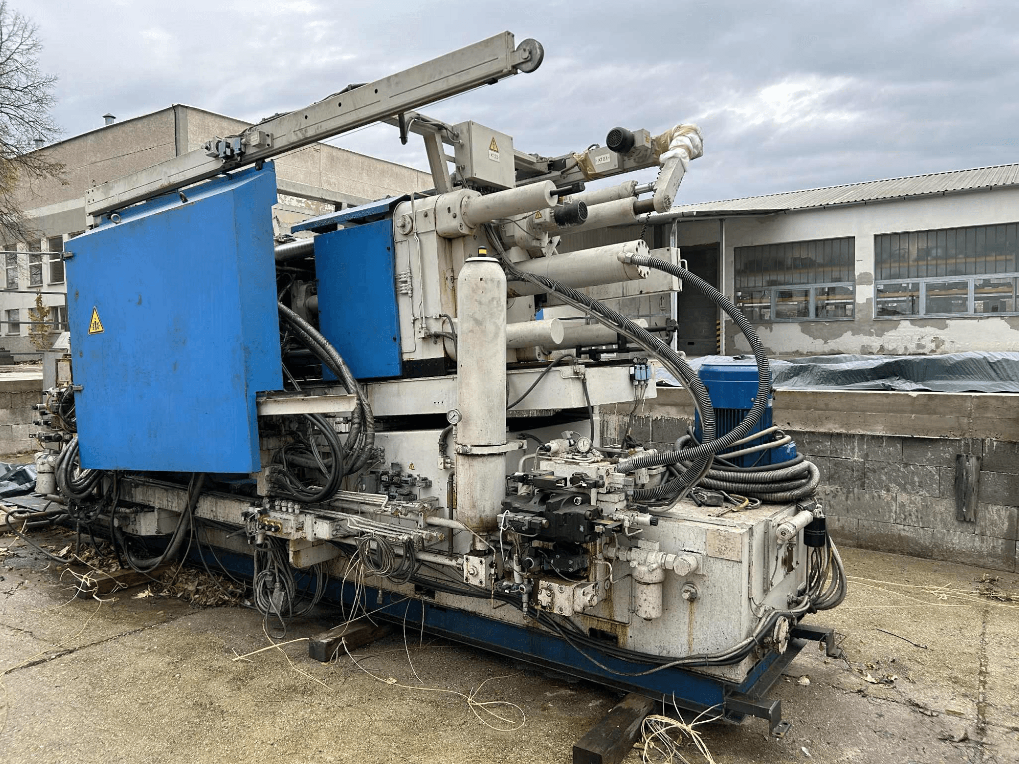 Stor hydraulisk industrimaskin med blått hölje, olika slangar och metallkomponenter, sedd från sidan.