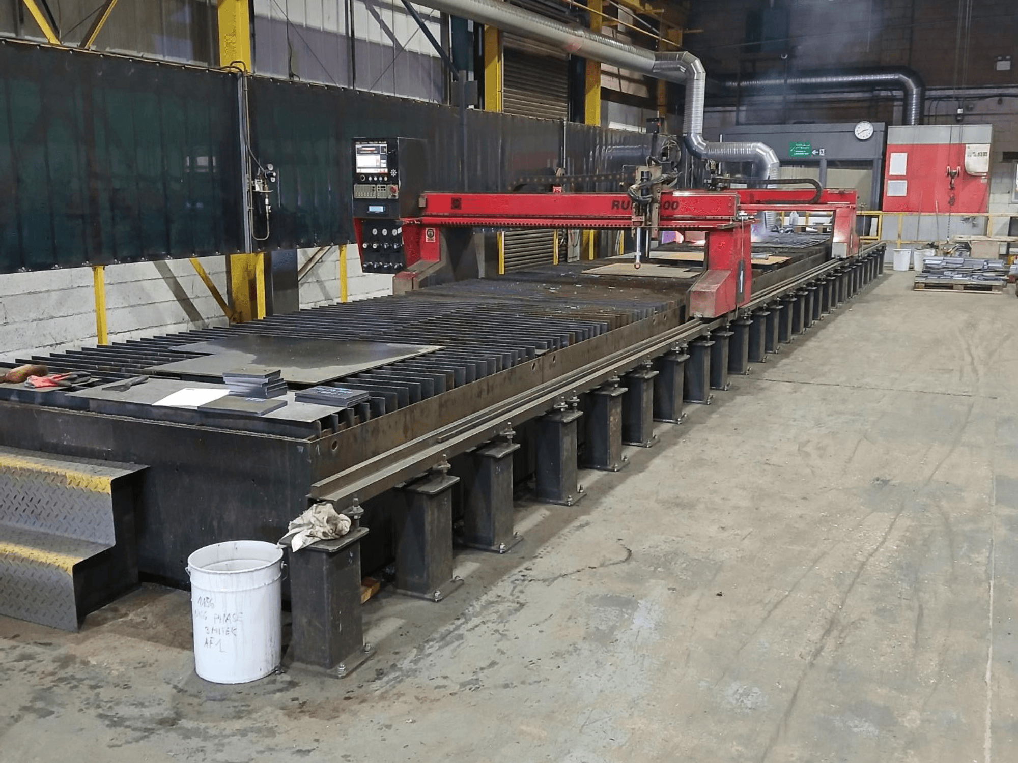 Röd RUIDA 300 skärmaskin, vy från sidan, med metallplåt på ett stålbord, placerad i ett industriellt arbetsutrymme.