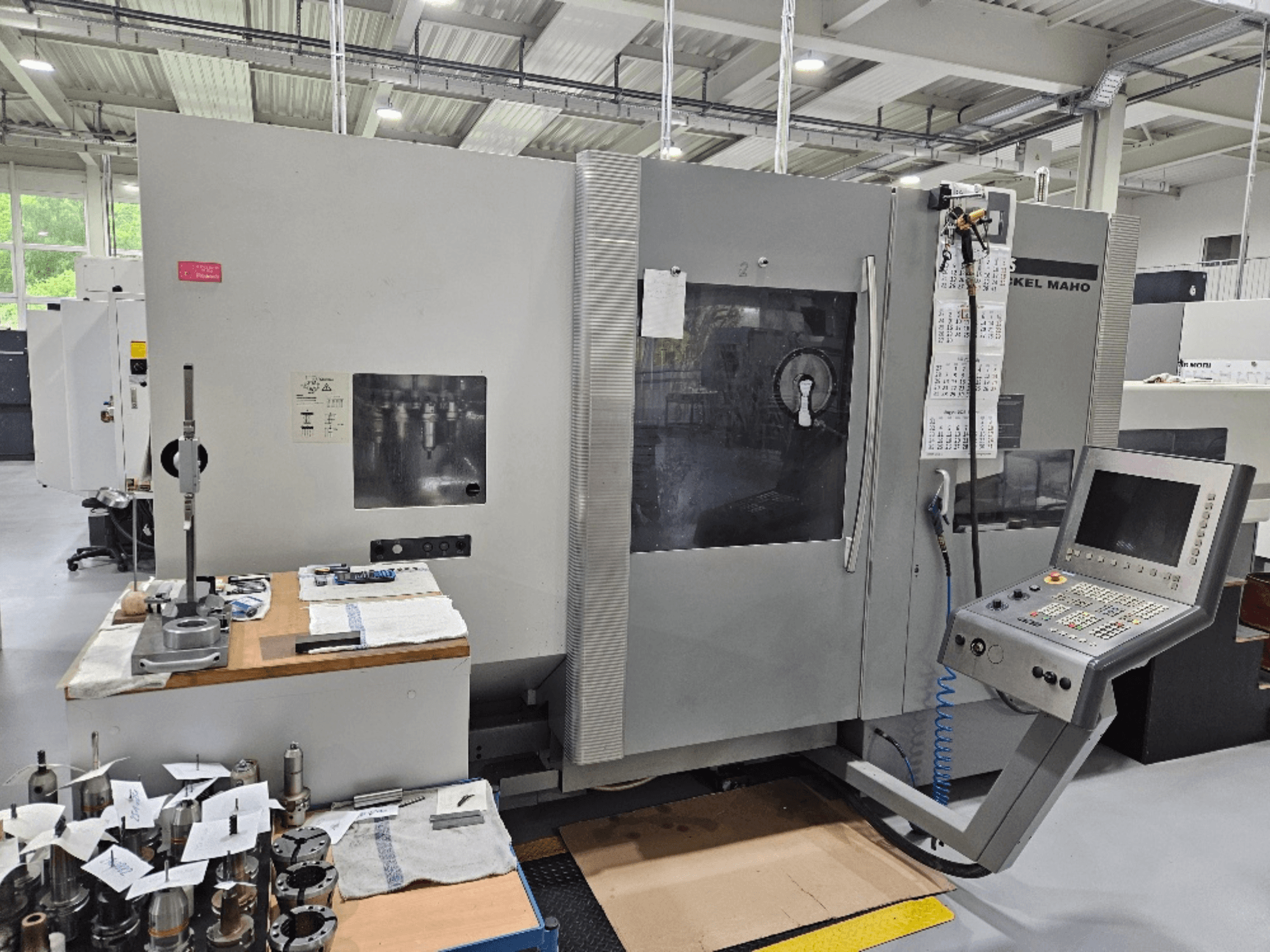 En vy framifrån av en DMG Mori DMU 70-maskin med kontrollpanel, verktyg på ett träbord och olika arbetsstycken i närheten.