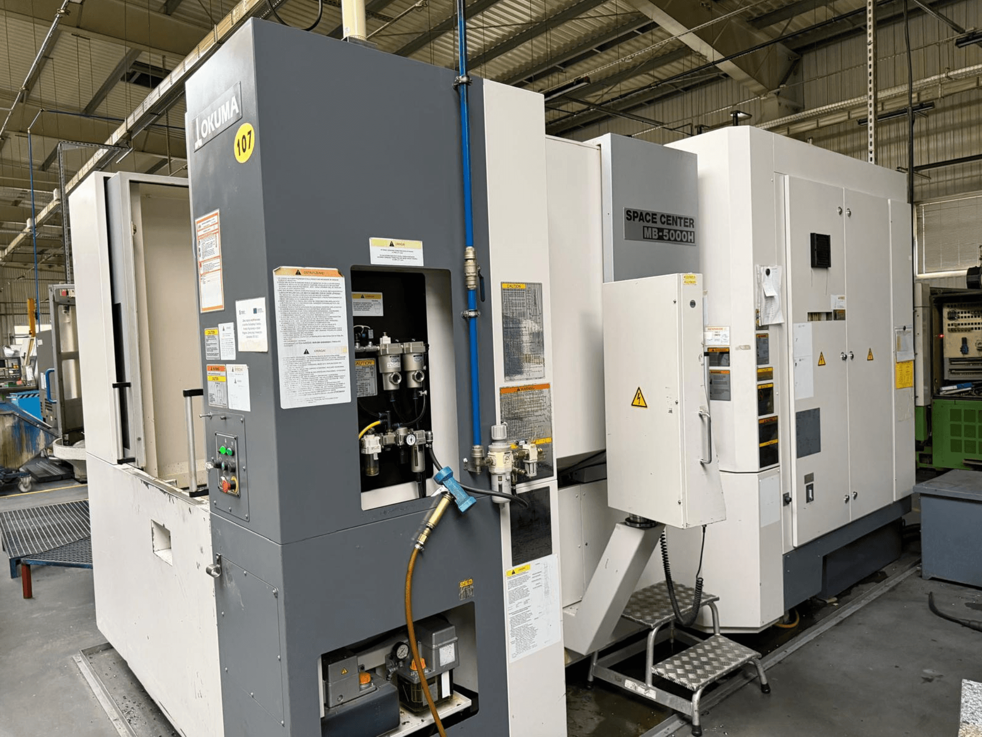 Okuma MB 5000H horisontell fleroperationsmaskin, sidovy som visar kontrollpanelen och maskinkomponenterna i en fabriksmiljö.