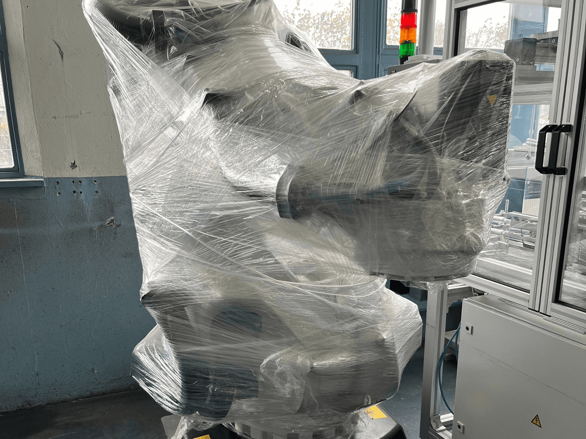 Industriell robotarm inlindad i plast, framifrån. Innehåller kontrollenhet och indikatorlampa. Varumärke ej synligt.