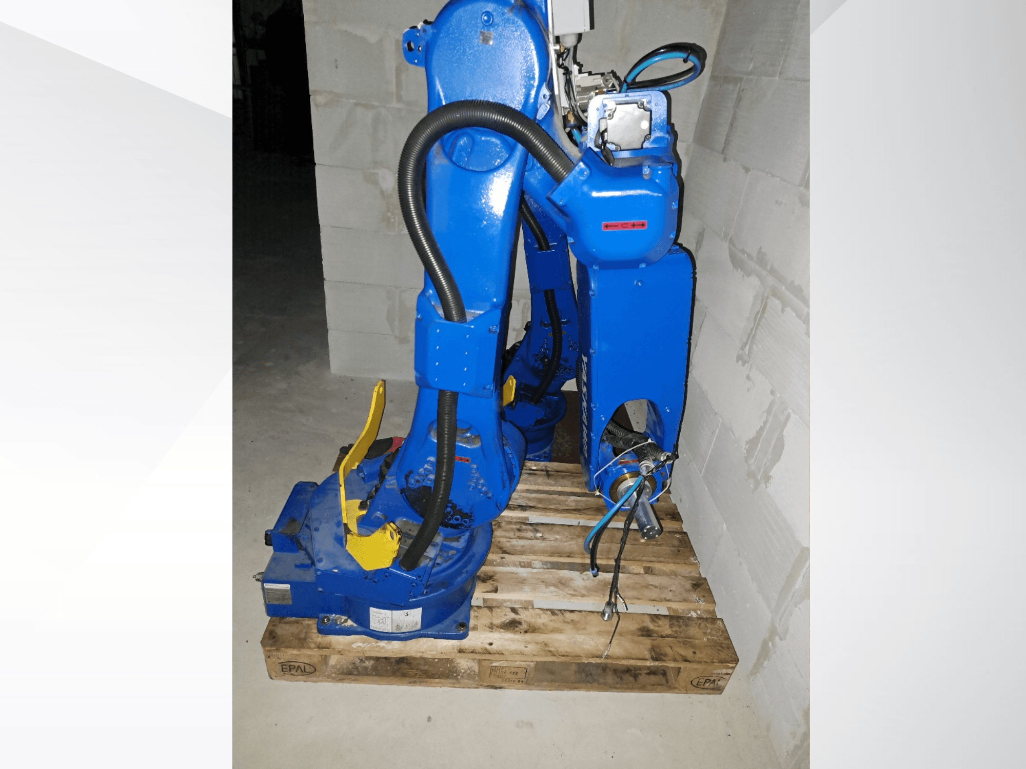 En Yaskawa Motoman GP25-robotarm i blått, sedd från sidan på en träpall, med kablar och mekaniska leder.