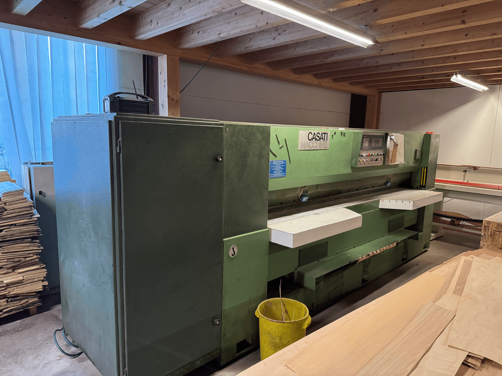 Grön CASATI träbearbetningsmaskin i en verkstad, framifrån, med kontrollpanel och vita arbetsytor, omgiven av trähögar.