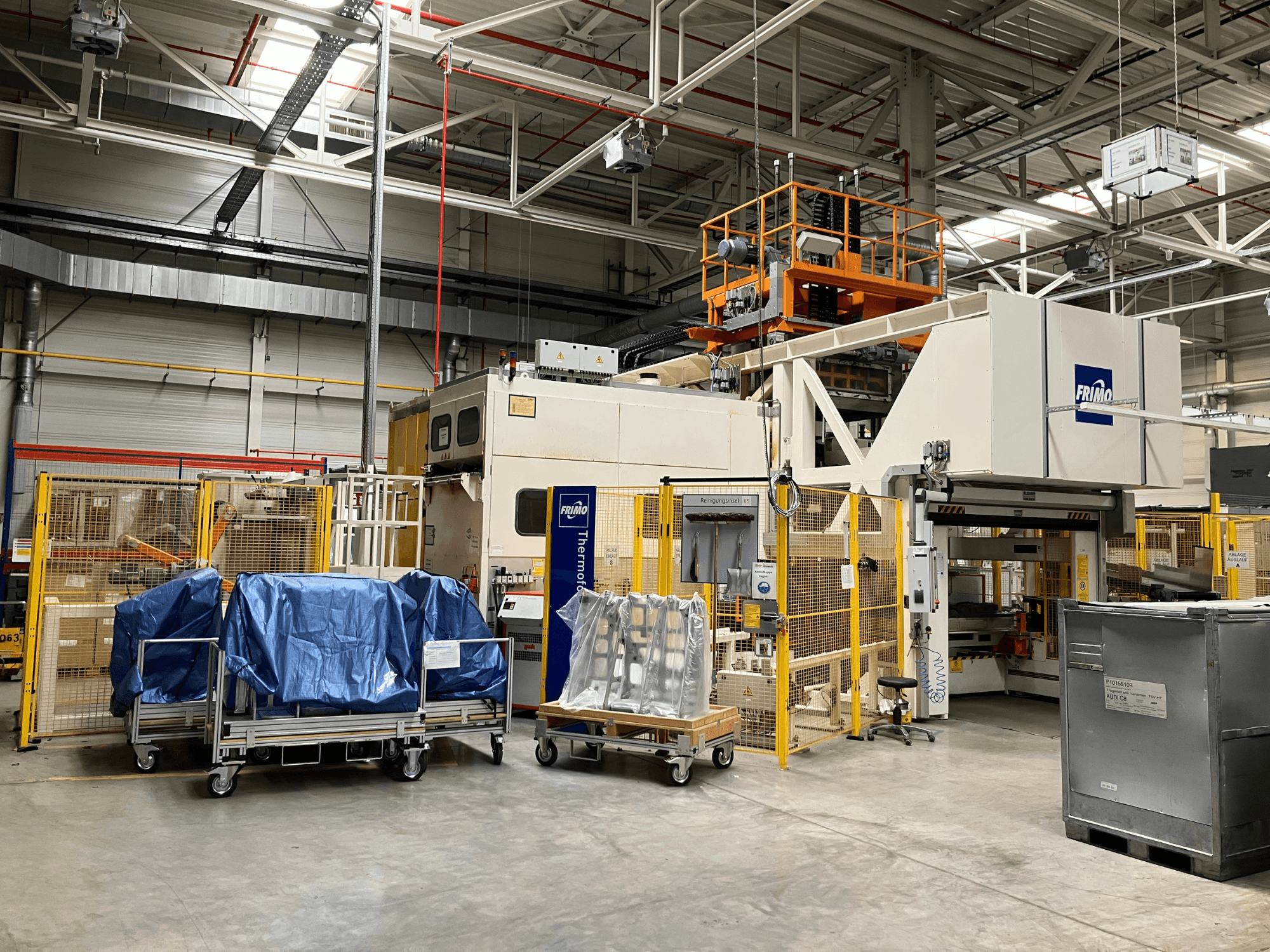 FRIMO FTL 80/200 Fullflex plastbearbetningsmaskin i en fabrik, vy framifrån med blå presenningar och säkerhetsbarriärer synliga.