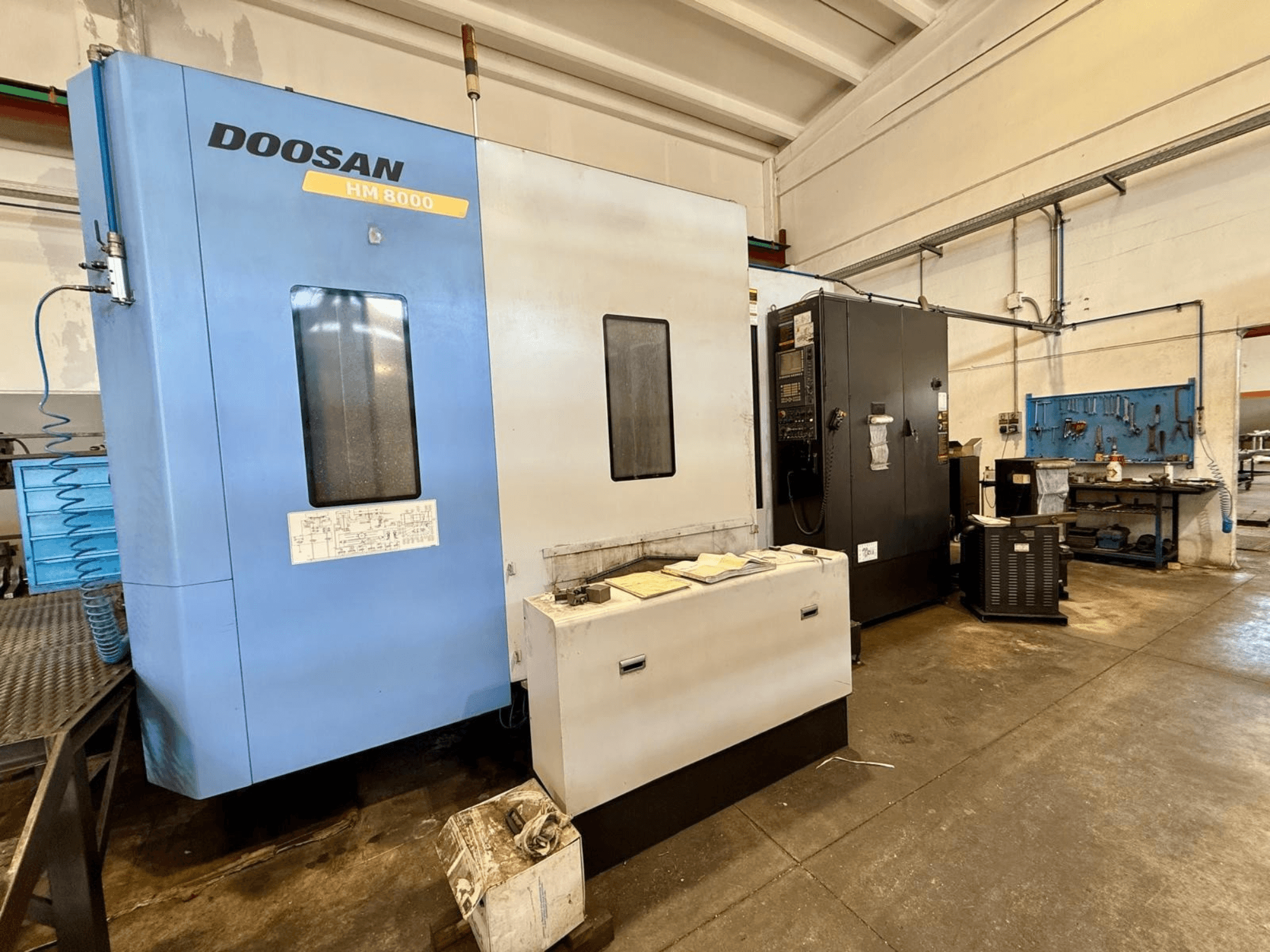 DOOSAN HM 8000 horisontellt bearbetningscenter i blått, framifrån, med kontrollpanelen och närliggande verkstadsutrustning.