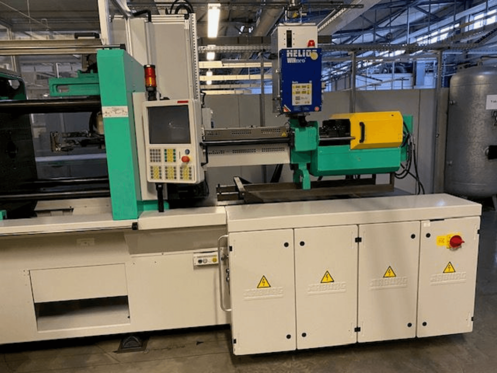 Industriell formsprutningsmaskin, modell HELIO, framifrån, med kontrollpanel, säkerhetsskydd och nödstoppsknapp.
