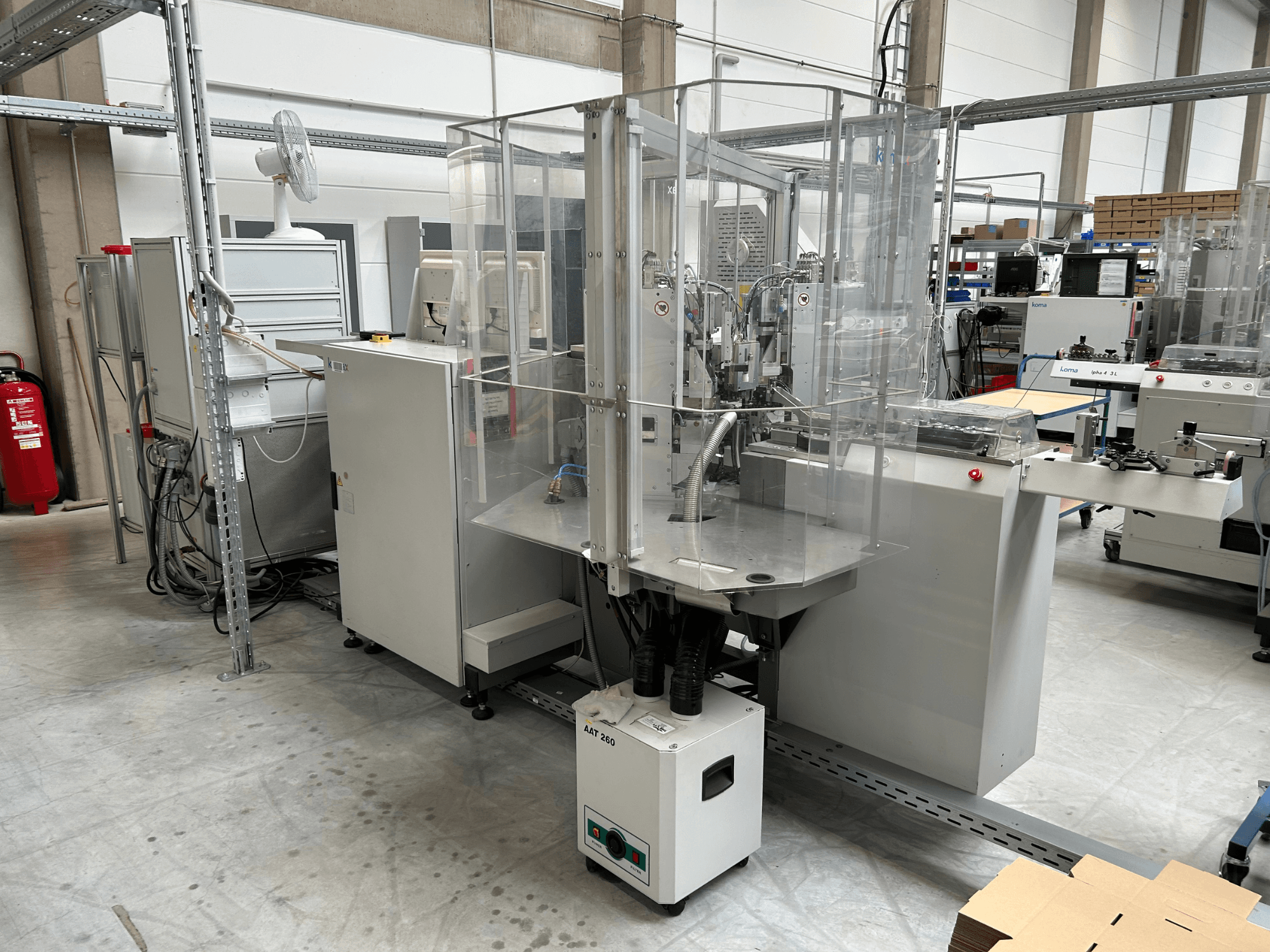 Automatiseringsutrustning av typen KOMAX Alpha 433 L i en fabriksmiljö. Utrustningen har ett genomskinligt skyddshölje och olika kontrollpaneler.