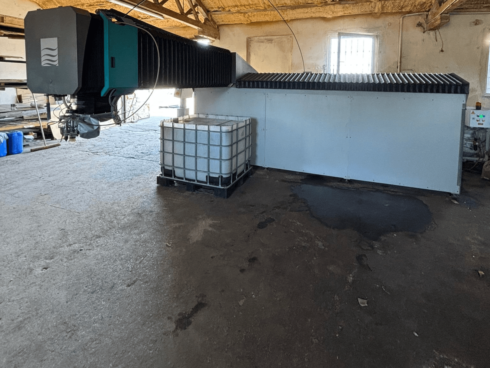 Industriell vattenjetmaskin i en verkstad, vy från sidan, med en stor vattentank och kontrollpanel synlig bredvid.