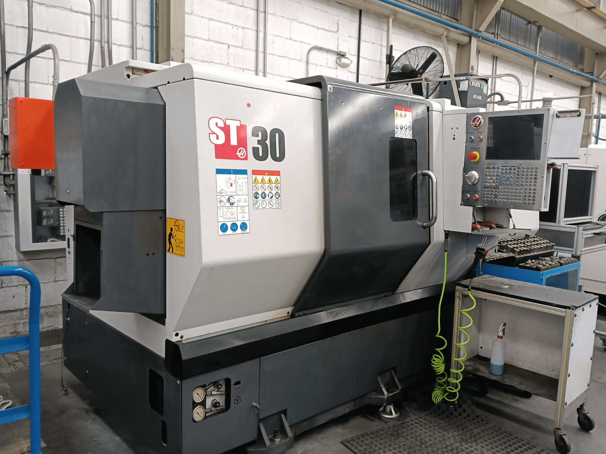 Alternativtext: Framifrånvy av den horisontella svarven HAAS ST-30 med kontrollpanel, säkerhetsetiketter och verktyg på en vagn i närheten.
