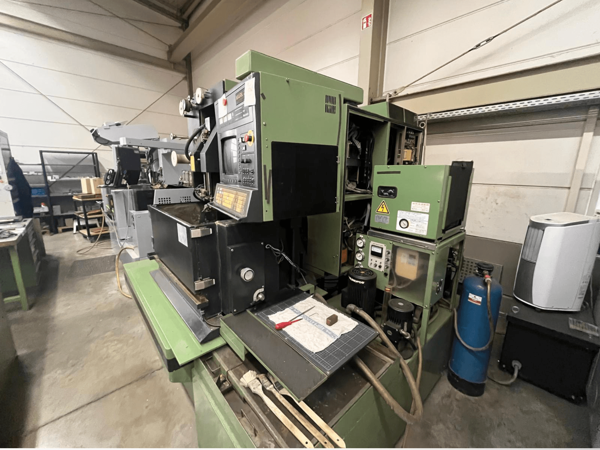 Grön industrimaskin med kontrollpanel, vy från sidan; inkluderar hydrauliska komponenter och arbetsyta med verktyg.