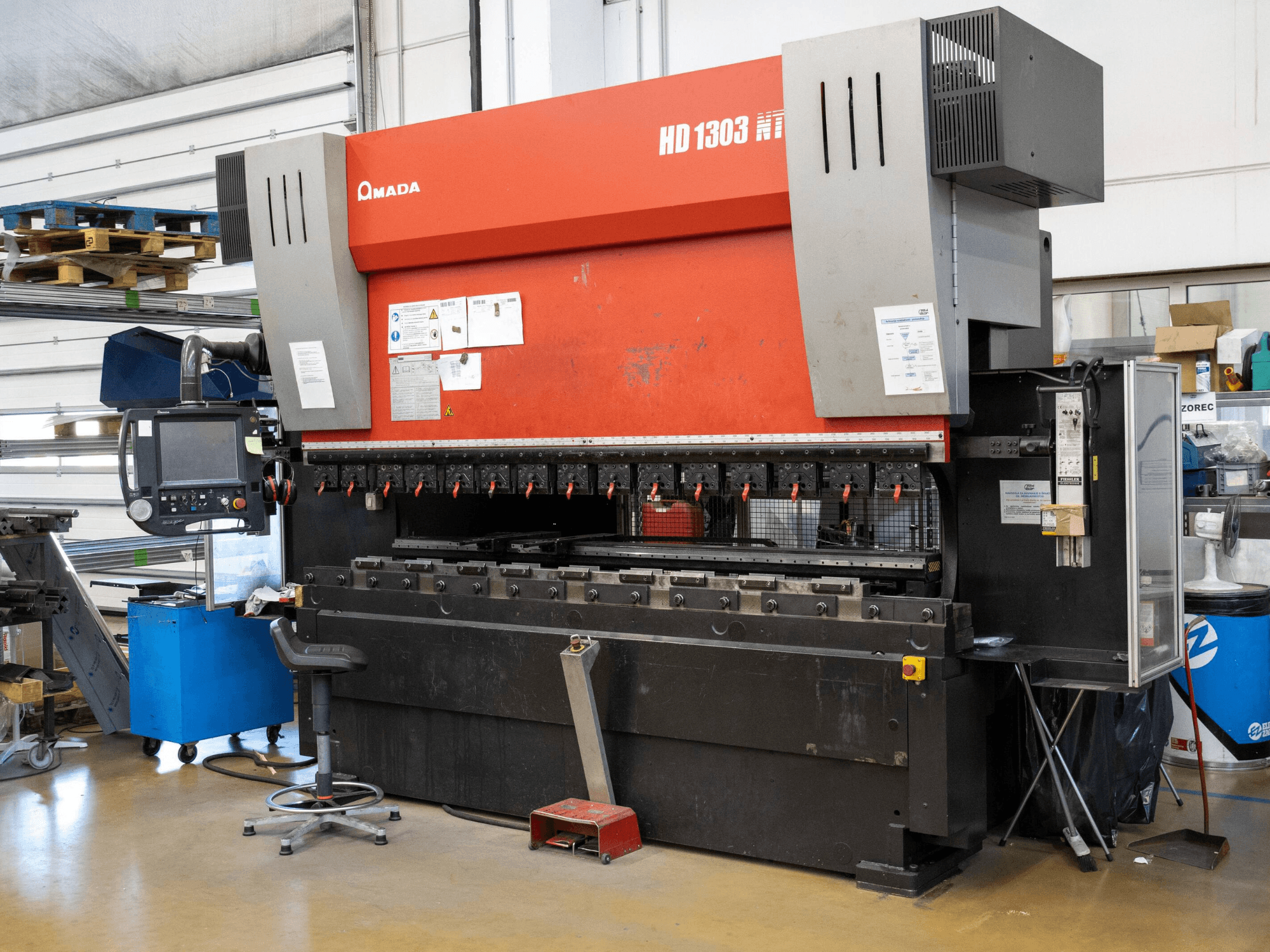 Amada HD 1303 hydraulisk kantpress i en verkstad, framifrån; har flera klämmor, kontrollpanel och justerbar pall.