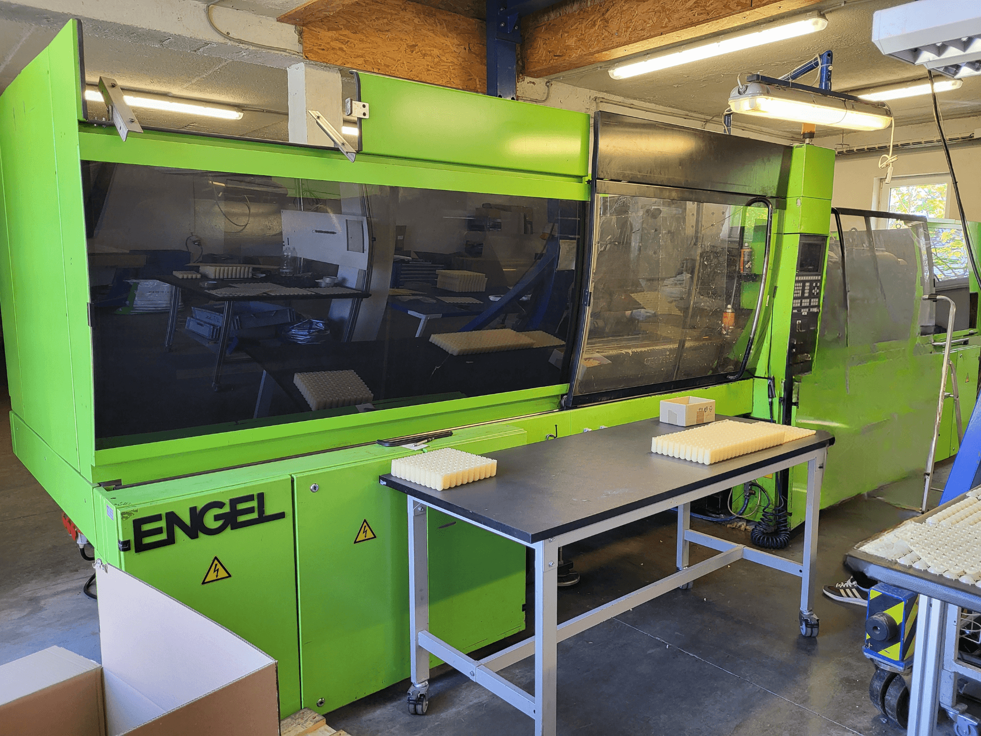 En hydraulisk formsprutningsmaskin av typen Green Engel ES1800/300HL sedd framifrån, med staplat material på ett arbetsbord.