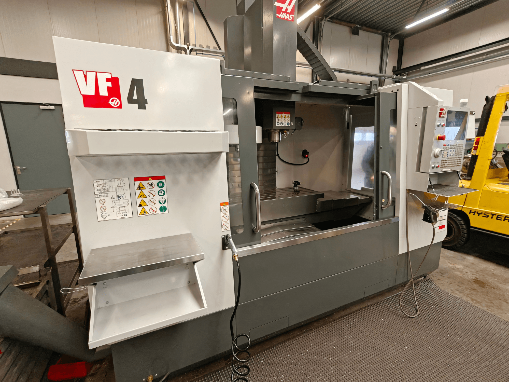 HAAS VF-4 vertikalt bearbetningscenter, framifrån, med kontrollpanel, arbetsområde och säkerhetsetiketter på sidan.