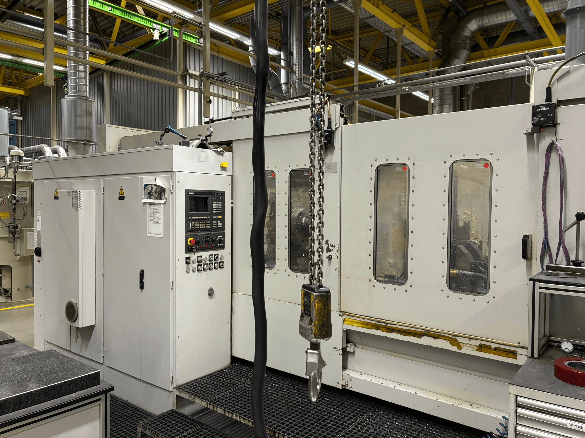 Industriell CNC-maskin med kontrollpanel, vy från sidan; har fönster, kedjor och metallkomponenter i en fabriksmiljö.