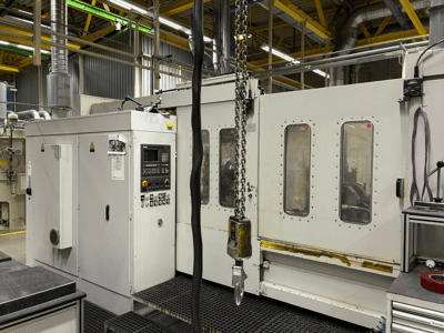 Industriell CNC-maskin med kontrollpanel, vy från sidan; har fönster, kedjor och metallkomponenter i en fabriksmiljö.