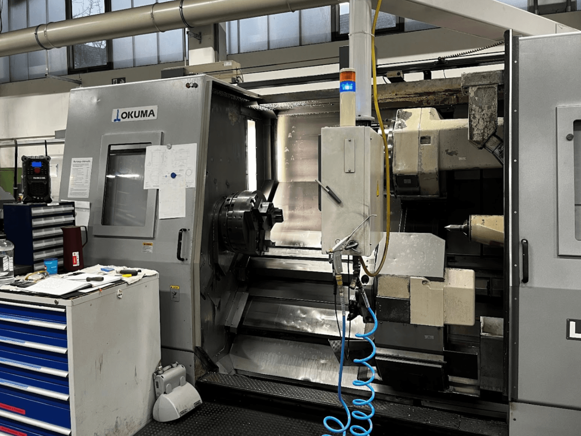 Okuma LU35-M horisontell svarv, vy framifrån. Utrustad med kontrollpanel, verktyg och stor chuck för bearbetningsoperationer.